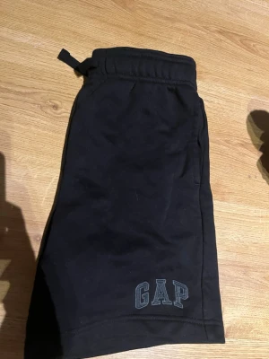 Svarta shorts från GAP med snörning - Svarta shorts från GAP med elastisk midja och snörning. GAP-logga broderad framtill. Tillverkade av mjuk bomullsmix, perfekta för avslappnade dagar. Klassisk och enkel design som funkar till det mesta. storleken är i barn storlek alltså inte vuxen storlek det innebär att dom är inte så stor som man tror att XXL är. 