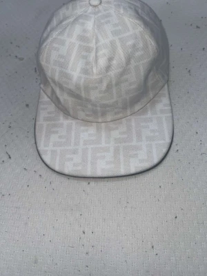 Vit keps från Fendi med monogram - Snygg vit keps från Fendi med det klassiska FF-monogrammet över hela kepsen. Kepsen har böjd skärm och är tillverkad i ett tyg som känns lyxigt och stilrent. Perfekt accessoar för att lyfta din outfit.