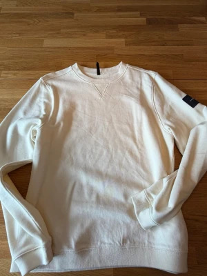 Biege sweatshirt från Sail Racing - Klassisk vit sweatshirt från Sail Racing med rund halsringning och ribbade muddar. Tröjan har en diskret logotyp-patch på ärmen och är tillverkad i mjukt bomullsmaterial. Perfekt för en clean och stilren look.aldrig använd.