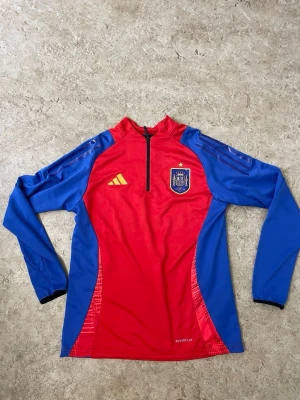 Spansk landslagströja Adidas röd/blå - Snygg långärmad träningströja från Adidas i rött och blått med Spaniens landslagsemblem på bröstet. Tröjan har halv dragkedja, gula detaljer och är tillverkad i lätt, ventilerande material med Aeroready-teknologi. Perfekt för fotbollsträning eller sport.