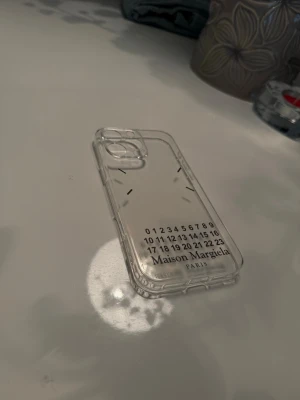 Maison Margiela genomskinligt mobilskal - Snyggt och stilrent genomskinligt mobilskal från Maison Margiela. Skalet är gjort i tålig plast och har det ikoniska numrerade trycket samt loggan på baksidan. Passar perfekt för dig som vill ha en clean look men ändå sticka ut med ett designerplagg till mobilen.
