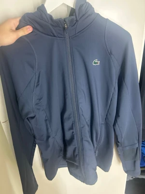 Marinblå Lacoste sport kofta - Snygg blå hoodie från Lacoste med dragkedja framtill och klassisk krokodillogga på bröstet. Tröjan har huva och är tillverkad i ett mjukt, stretchigt material som känns sportigt och bekvämt. Perfekt för en avslappnad stil.