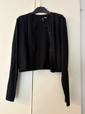 Svart kofta från H&M - En klassisk svart kofta från H&M med rund halsringning och knäppning framtill. Koftan har långa ärmar och är tillverkad i ett mjukt, tunt material som passar perfekt till lager-på-lager-stil.