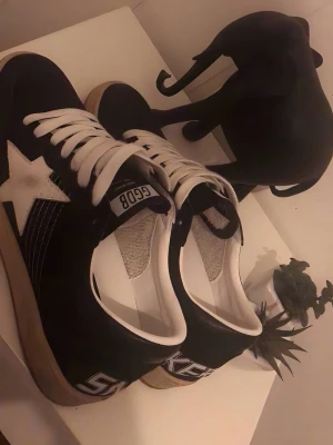 Golden Goose Ballstar sneakers svart/vit - Snygga Golden Goose Ballstar sneakers i svart mocka med vit stjärndetalj på sidan och vita skosnören. Skon har en retroinspirerad look med kontrasterande detaljer och texten 'SNEAKERS' på hälen. Insidan är vit och sulan har en vintagekänsla.
