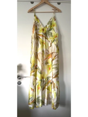 H&M - Lång blommig sommarklänning - storlek S - Härlig lång H&M klänning i storlek S med ett livfullt mönster av blad i olika nyanser. Klänningen har smala, justerbara axelband och en V-ringning. Midjan är markerad med dragskor som knyts i sidorna, vilket ger en smickrande passform. Kjolen är vid och avslutas med en volang nedtill, vilket skapar en fin rörelse. Finvävt material i 89% viskos, så bra material som andas. Perfekt för varma dagar och kan stylas för både avslappnade och mer uppklädda tillfällen. Mycket gott skick, som ny!