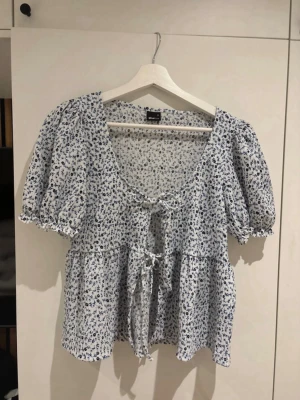 Blommig blus med puffärm från Gina Tricot - Supersöt blus från Monki i vitt med småblommigt blått mönster. Blusen har puffiga korta ärmar, två knytband framtill och en luftig, flowy passform. Perfekt till jeans eller kjol för en chill och trendig look. Materialet känns lätt och skönt mot huden.