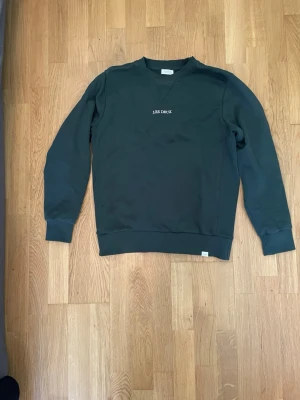 Mörkgrön sweatshirt från Les Deux - Snygg mörkgrön sweatshirt från Les Deux med diskret logga broderad på bröstet. Tröjan har rund halsringning, långa ärmar och ribbade muddar vid ärmslut och nederkant. Perfekt för en avslappnad och stilren look.