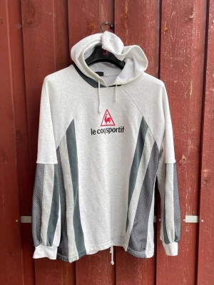 Grå hoodie från Le Coq Sportif - Snygg ljusgrå hoodie från Le Coq Sportif med svart och vit meshdetaljer längs ärmarna och logga på bröstet. Tröjan har huva med dragsko och avslappnad passform.