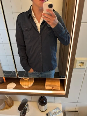 Mörkblå skjorta från Ben Sherman - Säljer en mörkblå skjorta från Ben Sherman med två bröstfickor och axelklaffar. Skjortan har knappar framtill och är långärmad. Perfekt till jeans för en clean och stilren look.