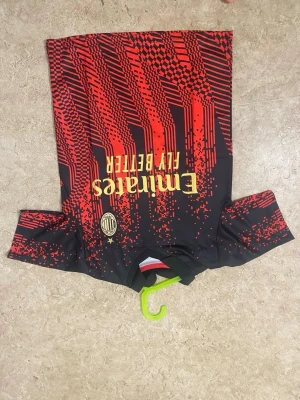 AC Milan matchtröja svart/röd - Snygg AC Milan matchtröja i svart med röda grafiska mönster och gula detaljer. Tröjan har korta ärmar, rund hals och klubbmärke på bröstet. Tillverkad i ett lätt och funktionellt material som passar perfekt till fotbollsträning eller match.