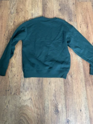 Mörkgrön sweatshirt från H&M - En enkel och stilren mörkgrön sweatshirt från H&M i loose fit. Tröjan har rund halsringning, långa ärmar och ribbade muddar vid ärmslut och nederkant. Perfekt för en avslappnad look och lätt att matcha med jeans eller joggers.
