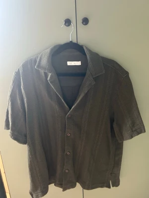 Olivgrön kortärmad skjorta från Zara - Säljer en olivgrön kortärmad skjorta från Zara med klassisk krage och knappar framtill. Skjortan är tillverkad i ett stickat material som ger en avslappnad och trendig look. Perfekt för dig som gillar stilrena plagg med lite struktur.