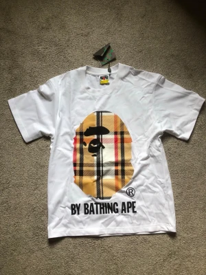 Vit BAPE t-shirt med rutigt tryck - Säljer en clean vit t-shirt från BAPE med det klassiska apansiktet i stort rutigt mönster på bröstet och texten 'BY BATHING APE' under.