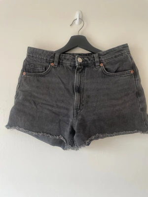 Svarta/gråa högmidjade jeansshorts - Snygga svarta högmidjade jeansshorts. Shortsen har knapp och dragkedja framtill samt bälteshällor. Perfekta för en avslappnad och trendig sommarlook. Storlek 28 från Monki.