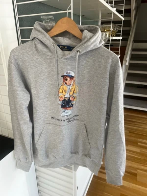 Grå hoodie Polo Bear Ralph Lauren - Säljer en grå hoodie från Polo Ralph Lauren med den ikoniska Polo Bear-trycket på bröstet. Hoodien har huva med dragsko, magficka och ribbade muddar. Perfekt för dig som gillar streetwear och klassisk stil. Materialet är mjukt och skönt bomullsblandat tyg. Storlek M
