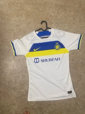 Vit Nike fotbollströja Al Nassr - Snygg vit fotbollströja från Nike med Al Nassr-logga på bröstet. Tröjan har blå och gula detaljer, korta ärmar med randiga muddar och tryck framtill. Tillverkad i lätt och ventilerande material, perfekt för träning eller match.