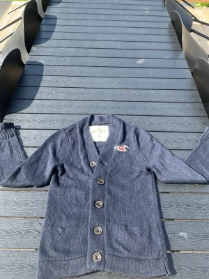 Mörkblå kofta från Hollister - Snygg mörkblå kofta från Hollister med v-ringning och fem knappar framtill. Koftan har ribbade muddar, två fickor och den klassiska Hollister-loggan broderad på bröstet. Perfekt för lager på lager och chill stil.