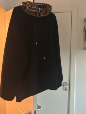 Svart hoodie med rutmönstrad huva - Säljer en svart hoodie med en riktigt snygg huva i klassiskt beige, svart och röd rutmönster. Hoodien har dragsko i huvan och en avslappnad passform. Perfekt för dig som vill ha en stilren men ändå unik look. Strl XL men sitter som en L