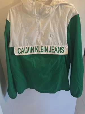 Grön och vit vindjacka Calvin Klein - Snygg vindjacka från Calvin Klein Jeans i grönt och vitt med stor logga framtill. Jackan har huva med dragsko, halv dragkedja och elastiska muddar. Perfekt för dig som gillar streetwear och vill sticka ut med en sportig look.