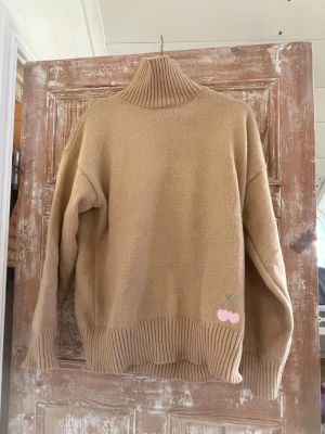 Beige polotröja Meet Me There - Stickad beige polotröja från Meet Me There x Filippa, med hög ribbad krage och breda muddar. Tröjan har en liten broderad rosa körsbärsdetalj nedtill. Perfekt för dig som gillar en clean och mysig stil.