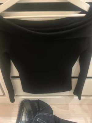 Svart offshoulder topp från 157 - Svart långärmad offshoulder topp från 157. Snygg och tight passform som framhäver axlarna. Perfekt att styla med jeans eller kjol för en trendig look. Mjuk och stretchig känsla i materialet.