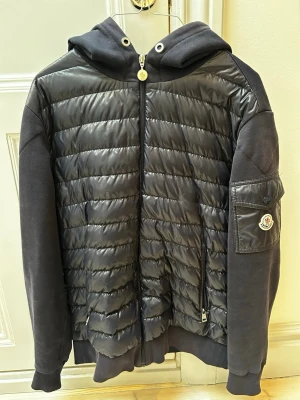 Moncler cardigan - Snygg svart cardigan från Moncler med huva. Jackans snören har jag lyckats tappa bort samt moncler lappen i jackan har lossnat men min skräddare har sytt fast igen. Därav det låga priset.