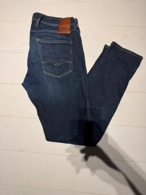 Mörkblå jeans från Replay - Snygga mörkblå jeans från Replay med klassisk femficksmodell och diskreta slitningar framtill. Jeansen har raka ben och detaljerade sömmar på bakfickorna samt en läderpatch med logga i midjan. Perfekta för en avslappnad och trendig look.