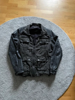 Mörkgrön jacka med skinnsärmar - Snygg mörkgrön jacka med detaljer i fuskskinn på ärmarna och axlarna. Jackan har flera fickor med dragkedja och knappar framtill samt en hög krage. Materialmixen ger ett coolt intryck och passar perfekt till streetwear-stilen.