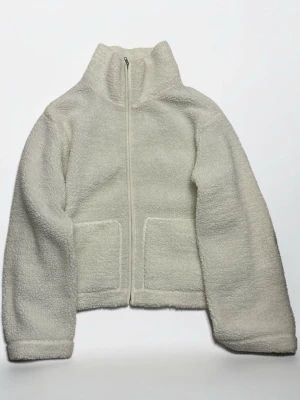 Vit teddyjacka fleece med dragkedja – oversized – stl M - Mysig vit teddyjacka med hög krage och hel dragkedja framtill. Jackan har två stora fickor på framsidan och är gjord i fluffigt, mjukt material som ger en avslappnad och trendig look. Perfekt för kyliga dagar och enkel att matcha med olika outfits.