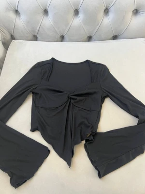Svart långärmad topp med twist - Trendig svart långärmad topp med twistad detalj framtill och asymmetrisk nederkant. Toppen har vida ärmar och är croppad, vilket ger en edgy och modern look. Perfekt att styla med höga jeans eller kjol.