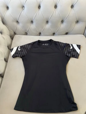 Nike dri-fit t-shirt  - Snygg svart sporttröja från Nike med korta ärmar. Ärmarna har ett grått och svart mönster samt vita Nike-loggor. Tillverkad i ett lätt och snabbtorkande material som passar perfekt till träning eller sportaktiviteter.