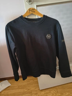 Svart sweatshirt från Philipp Plein - Cool svart sweatshirt från Philipp Plein med logga på bröstet och ett band med märkesnamnet på ärmen. Tröjan har rund halsringning och långa ärmar. Perfekt för dig som gillar streetwear och vill ha något stilrent men ändå exklusivt. Står att den är M i storlek men den passar som en S, därför jag säljer den, pris går att diskutera 