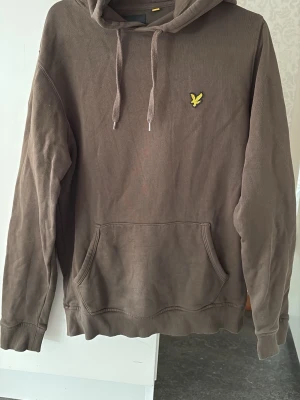 Mossgrön hoodie från Lyle & Scott - Säljer en mossgrön hoodie från Lyle & Scott med klassisk gul logga på bröstet. Hoodien har huva med dragsko och en stor magficka framtill. Tillverkad i mjukt bomullsmaterial och har ribbade muddar vid ärmslut och nederkant. Perfekt för en avslappnad stil. Mycket fint skick!