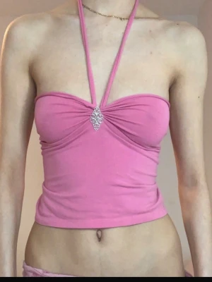 Rosa bandeau topp med knyt och smycke - Trendig rosa bandeau topp med justerbara knytband som kan bäras på olika sätt. Toppen har ett dekorativt smycke framtill och är tillverkad i mjukt bomullsmaterial. Perfekt för dig som vill ha en unik och snygg look.