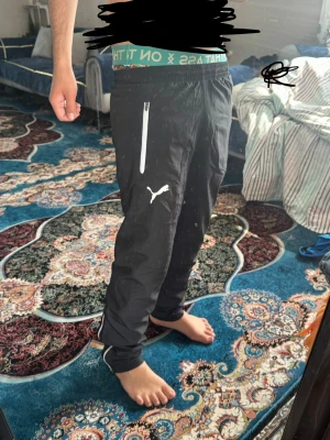 Svarta mjukisbyxor från Puma - Säljer ett par svarta mjukisbyxor från Puma med vit logga på benet och dragkedjeförsedd ficka. Byxorna har smal passform och resår i midjan. Perfekta för chill eller träning, riktigt sköna och stilrena.