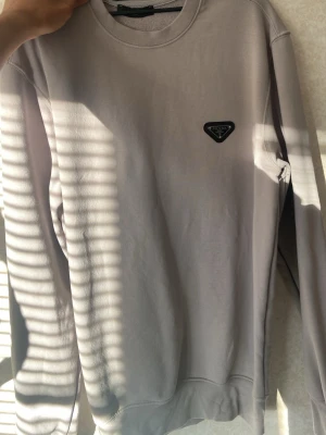 Ljusgrå sweatshirt från Prada - Minimalistisk ljusgrå sweatshirt från Prada med klassisk rund halsringning och lång ärm. Tröjan har en ikonisk svart Prada-logga i metall på bröstet. Tillverkad i mjukt bomullsmaterial för en clean och stilren look.