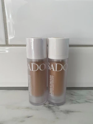 SADOER Make Up The Glow Foundation - Två flaskor SADOER Make Up The Glow Foundation i pumpflaska. Färgen är medium beige med varm underton. Flaskorna är genomskinliga med vita lock och text i vitt. Perfekt för en naturlig och fräsch bas.