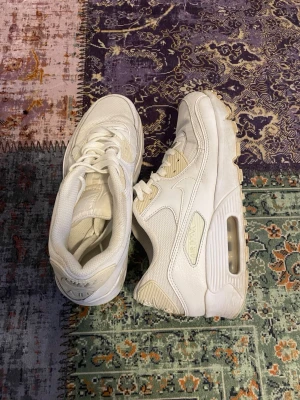Vita Nike Air Max 90 sneakers - Säljer ett par klassiska vita Nike Air Max 90 sneakers med synlig luftdämpning i sulan. Skorna har en chunky siluett, mesh och läderdetaljer samt vita skosnören. Perfekta för dig som gillar streetwear och sportig stil.