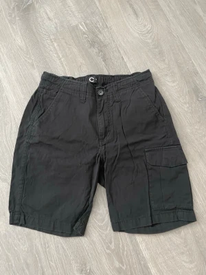 Svarta cargoshorts med fickor - Säljer ett par svarta cargoshorts med flera praktiska fickor och knappstängning fram. Shortsen har elastisk midja baktill för extra komfort och en avslappnad passform. Perfekta för varma dagar och enkel att matcha med olika stilar.