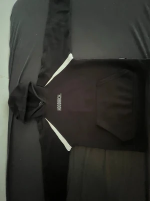 Svart HOODRICH hoodie med vita detaljer - Säljer en svart hoodie från HOODRICH med vita detaljer längs axlarna och bröstet. Stor ficka framtill och tydlig HOODRICH-logga tryckt på bröstet. Klassisk huva och mjukt material som känns skönt mot huden. Perfekt för dig som gillar streetwear.