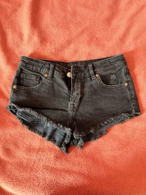 Svarta denimshorts - Säljer ett par svarta denimshorts med fransig nederkant. Shortsen har knapp och dragkedja framtill samt små slitningar på ena fickan. 