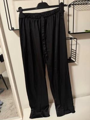 Svarta satinpajamasbyxor med dragsko - Snygga svarta pyjamasbyxor i satin med elastisk midja och dragsko för perfekt passform. Byxorna har raka ben och en mjuk, glansig finish som känns lyxig mot huden. Perfekta för mysiga hemmakvällar.