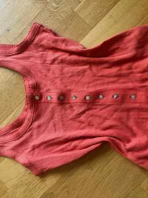 Ribbad korallfärgad linnetopp Next - Snygg ribbad linnetopp från Next i en fräsch korallröd nyans. Toppen är ärmlös och har dekorativa knappar framtill som ger en cool detalj. Perfekt att styla med jeans eller kjol för en trendig look.