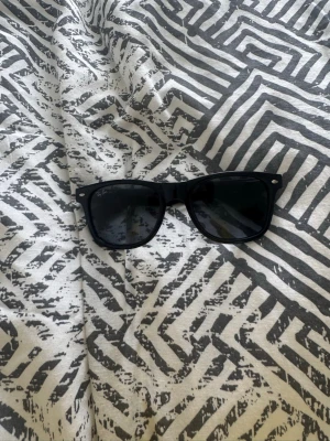 Ray-Ban Wayfarer solglasögon - Klassiska svarta Wayfarer solglasögon från Ray-Ban med tidlös design och mörka glas. Bågarna är i plast och har Ray-Ban-loggan på sidan. Perfekta för att höja din stil under soliga dagar.