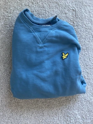 Blå sweatshirt från Lyle & Scott - En stilren blå sweatshirt från Lyle & Scott med klassisk rund halsringning och det ikoniska gula fågel-logotypen broderad på bröstet. Tröjan har långa ärmar och ribbade muddar vid ärmslut och nederkant. Perfekt för en avslappnad look.
