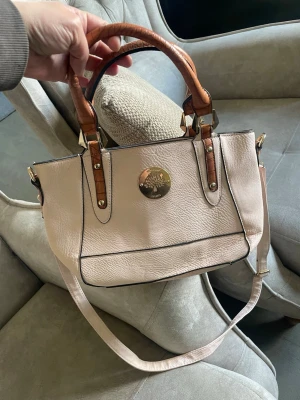 Ljusrosa handväska med bruna handtag - Snygg ljusrosa handväska från Mulberry med bruna handtag i croco-imitation och avtagbar axelrem. Väskan har en rund guldfärgad logga framtill och svarta kanter som ger en cool kontrast. Perfekt storlek för det viktigaste och passar till många olika outfits.