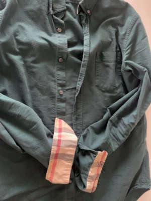 Grön skjorta med rutiga manschetter - Snygg grön skjorta från Burberry med klassiska rutiga detaljer på insidan av manschetterna och diskret broderad logga på bröstfickan. Skjortan har knappar framtill och är långärmad. Perfekt för dig som gillar stilrena plagg med en twist.