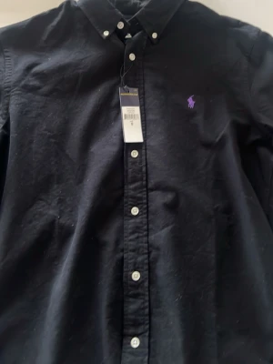 Svart skjorta från Ralph Lauren - Klassisk svart skjorta från Ralph Lauren med lila broderad logga på bröstet. Skjortan har knappar hela vägen fram och button-down krage. Tillverkad i bomull och har lång ärm. Perfekt för en clean och stilren look.