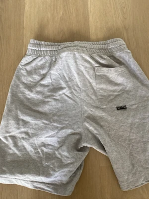 Grå sweatshorts med snörning - Säljer ett par grå sweatshorts med resårmidja och vit snörning. Shortsen har sidofickor och är tillverkade i mjuk bomull, perfekta för chill dagar eller träning. Enkel och avslappnad stil som passar till det mesta.