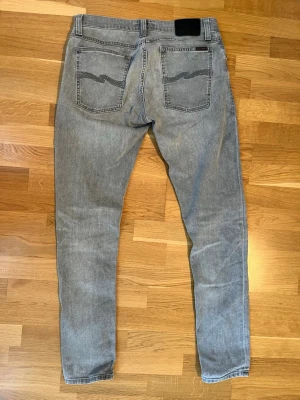 Grå slim fit jeans från Nudie Jeans - Säljer ett par grå slim fit jeans från Nudie Jeans med klassisk femficksmodell och svarta detaljer på bakfickorna. Jeansen har normal midja och är tillverkade i mjuk denim med stretch för extra komfort. Perfekta för en avslappnad och trendig look. Modell Thinn finn, storlek 34,32. 